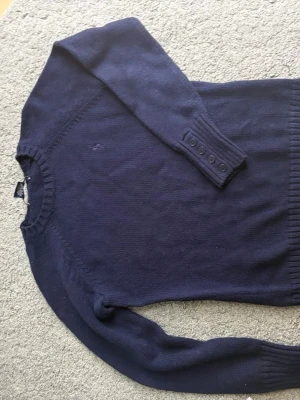 Mörkblå stickad tröja med knappar - Mörkblå stickad tröja med rund hals och långa ärmar. Ärmsluten har ribbstickning och fyra dekorativa knappar. Perfekt för lager på lager och enkel att matcha med jeans eller kjol.