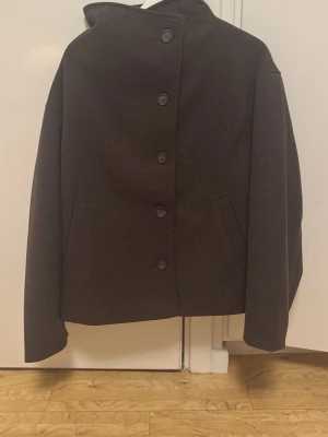 Mörkbrun kappa med hög krage - Stilren mörkbrun kappa med hög krage och fem knappar framtill. Kappan har raka ärmar och två snedställda fickor. Materialet ser ut att vara tjockt och mjukt, perfekt för kyligare dagar. Enkel och modern design som passar till det mesta.