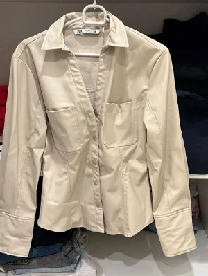 Beige skinnjacka från Zara - Snygg beige skinnjacka från Zara med klassisk krage, två bröstfickor och knäppning framtill. Figurnära passform och långa ärmar med knappdetaljer vid ärmslut. Perfekt för dig som vill ha en stilren och trendig look.