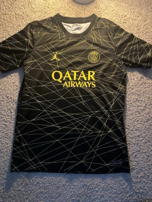 Svart PSG matchtröja Jordan x Qatar - Snygg svart PSG matchtröja i samarbete med Jordan och Qatar Airways. Tröjan har gula detaljer, PSG-logga på bröstet och ett unikt mönster av tunna gula linjer över hela plagget. Tillverkad i lätt och funktionellt material, perfekt för fotboll eller träning.