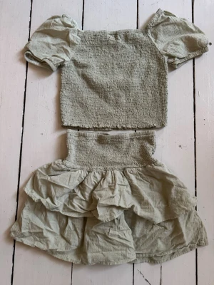 Ljusgrönt tvådelat set med puffärm - Säljer ett ljusgrönt tvådelat set med puffärmar och smockdetaljer. Toppen har korta puffärmar och smockad kropp, medan kjolen har bred smock i midjan och dubbla volanger. Setet är gjort i ett mjukt, tunt tyg som ger en avslappnad look.