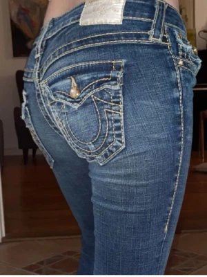 Blå True Religion jeans med kontrastsömmar och diamanter - Säljer ett par blå jeans från True Religion med ikoniska kontrastsömmar och stora fickor med dekorativ rhinestone knappdetalj. Jeansen har låg midja , en ficka saknar diamant 
