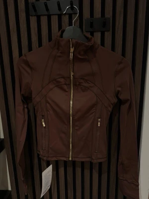 Brun jacka från Lululemon med dragkedja - Snygg brun jacka från Lululemon med guldig dragkedja framtill och två fickor med dragkedja. Köpt på en outlet. Jackan har en figurnära passform och hög krage, perfekt för en sportig och stilren look. Tog helt fel storlek vilket är därför jag säljer den. 