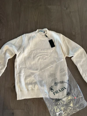 Vit stickad tröja från Prada - Snygg vit stickad tröja från Prada med diskret broderad logga och text framtill. Tröjan har rund halsringning, långa ärmar och ribbade muddar vid ärmslut och nederkant. Perfekt för dig som gillar exklusiv och stilren design.