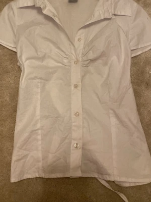 Vit blus från Gina Tricot - Snygg vit blus från Gina Tricot med korta ärmar och knappar framtill. Blusen har krage och lätt rynk vid bysten för en feminin touch. Storlek M men skulle säga att den sitter mer som en S/Xs. Tillverkad i ett tunt och mjukt tyg, perfekt för en fräsch och stilren look.