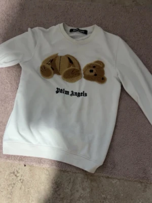 Vit stickad tröja soppar Palm Angels - Vit stickad tröja från Palm Angels med ett bro soppar nallemotiv i beige och brunt på bröstet samt svart logga under. Tröjan har rund halsringning och långa ärmar. Materialet känns mjukt och passar perfekt för dig som gillar streetwear och unika detaljer.