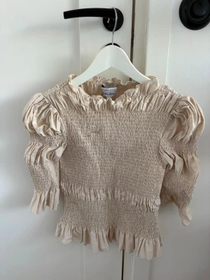 Beige smockad blus proppad med volanger - Supersöt beige blus med smockad kropp och puffiga långärmar. Volangdetaljer vid hals, evenly över ärmar och nederkant ger en romant tear och trend step. Perfekt för dig som gillar en feminin och chic stil. Materialet känns lätt och soft.