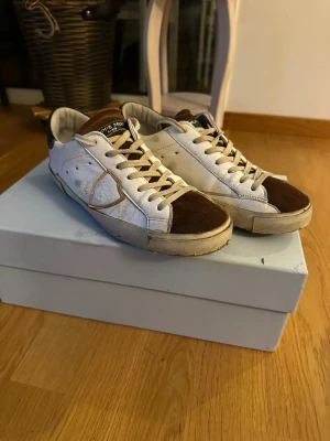 Philippe Model vita och bruna sneakers - Snygga vita sneakers från Philippe Model med bruna mockadetaljer på tå och häl. Klassisk låg modell med vita snören och kontrasterande sula. Märkets logga syns tydligt på sulan och sidan. Perfekta för en trendig streetwear-look.