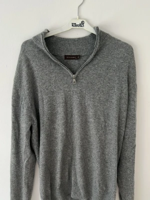 Grå stickad tröja med halvzip - Mysig grå stickad tröja från Oscar Jacobson med halvzip och hög krage. Tröjan har långa ärmar och ribbade muddar vid ärmslut och nederkant. Perfekt för lager på lager och enkel att matcha till olika outfits.