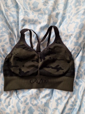 Camo sport-bh från Gavelo - Sport-bh i mörkgrönt camomönster från Gavelo. Bh:n har brottarrygg med dubbla axelband och bred resår under bysten för extra stöd. Tillverkad i stretchigt material som passar perfekt för träning och gym.