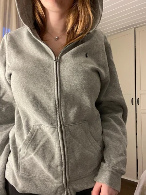 Grå zip hoodie från Polo Ralph Lauren - Säljer en grå hoodie med dragkedja från Polo Ralph Lauren. Klassisk modell med huva, fickor framtill och liten mörkblå logga på bröstet. Tillverkad i mjukt sweatshirtmaterial som är perfekt för chill dagar. Passar dig som gillar enkel och stilren streetwear. Hoodien känns som storlek L men är XL