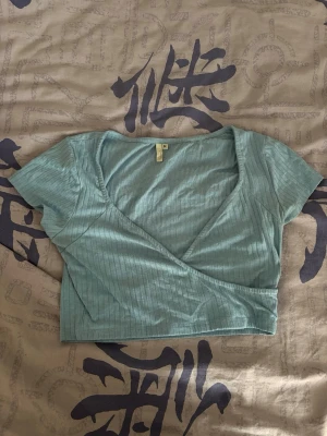 Ljusblå omlottopp med ribbad struktur - Ljusblå omlottopp med ribbad struktur och korta ärmar. Toppen har en croppad passform och djup v-ringning, Nelly.