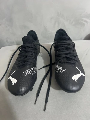 Svarta Puma fotbollsskor - Snygga svarta fotbollsskor från Puma Future-serien med vita detaljer och logga. Skorna har snörning, låg profil och ovandel i syntetmaterial med textur för bättre bollkontroll. Perfekta för dig som vill ha stilrena och funktionella skor på planen.