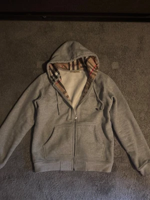Grå zip hoodie med Burberry-mönster - Säljer en grå hoodie med dragkedja från Burberry. Huvan är fodrad med det klassiska rutmönstret i beige, svart och rött. Hoodien har två fickor fram, dragsko i huvan och en diskret Burberry-logga broderad på bröstet. Perfekt för en chill och stilren look.