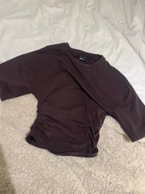 Brun crop top med rynkade sidor - Säljer en brun crop top från gina med rynkade detaljer på ena sidan. T-shirten har korta ärmar och rund halsringning. Materialet är mjukt och stretchigt, perfekt för en avslappnad och trendig look.