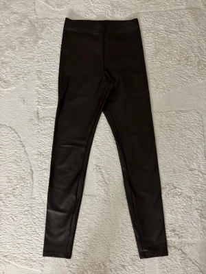 Svarta leggings i fuskskinn - Snygga svarta leggings i fuskskinn-look med hög midja och smal passform. Perfekta för att ge outfiten en edgy vibe. Stretchigt material som sitter tajt och bekvämt. Inga synliga fickor eller dragkedjor.