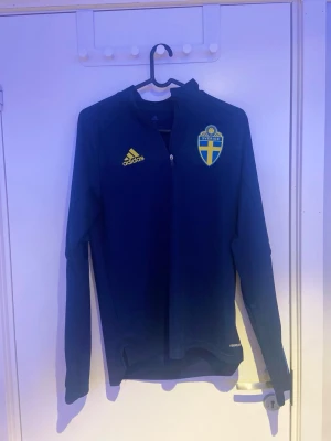 Svenska landslaget Adidas träningströja - Mörkblå långärmad träningströja från Adidas med Sveriges landslagsemblem på bröstet och gula Adidas-loggan. Dragkedja framtill och färgade ränder på ärmarna, blå på ena och gula på andra. Perfekt för fotbollsträning eller support.
