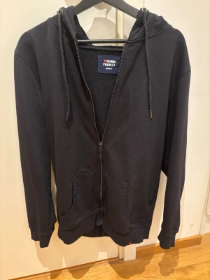 Svart zip-hoodie från Denim Project - Svart hoodie med dragkedja från Denim Project. Klassisk modell med huva och dragsnören, två fickor framtill och ribbade muddar vid ärmslut och nederkant. Perfekt för en avslappnad stil och enkel att matcha med andra plagg.