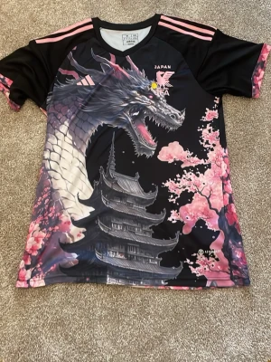 Japan Adidas topp med drakmotiv - Unik svart och rosa topp från Adidas med Japan-tema. Framsidan har ett stort drakmotiv, körsbärsblommor och ett traditionellt japanskt tempel. Korta ärmar med rosa ränder på axlarna och detaljerad tryckt design. Perfekt för dig som gillar streetwear och coola prints