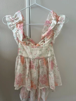 Blommig volangblus med breda axelband - Söt blommig blus med volanger och breda axelband. Toppen är i ljusa toner med rosa och orange blommor, har en luftig passform och är tillverkad i ett tunt, mjukt material. Perfekt för varma dagar och ger en romantisk vibe. Använd en gång 