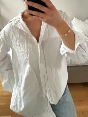 Vit oversized skjorta i bomull - Klassisk vit skjorta i bomull med avslappnad, oversized passform. Skjortan har långa ärmar, knappar framtill och en mjuk, lite skrynklig struktur. Perfekt att styla med jeans eller kjol för en enkel och trendig look.