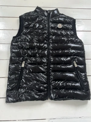 Svart dunväst från Moncler - Snygg svart dunväst från Moncler med glansig finish och ståkrage. Västen har två dragkedjeförsedda fickor framtill och Moncler-logga på bröstet. Perfekt att slänga över en hoodie eller tröja för extra värme. Klassisk quiltad design och lätt vaddering. Dock inte original 