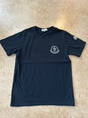 Svart Moncler t-shirt med logga - Svart t-shirt från Moncler med rund halsringning och vit Moncler-logga på bröstet. Klassisk passform och korta ärmar. Liten Moncler-patch på vänster ärm. Tillverkad i mjuk bomull. Storlek small passar någon med storlek XS. 