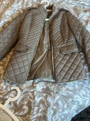 Beige quiltad jacka med svarta detaljer - Snygg beige quiltad jacka med stora fickor och svarta armbågslappar. Jackan har dragkedja framtill, hög krage och axelklaffar för en cool look. Perfekt för dig som gillar stilrena och praktiska plagg.