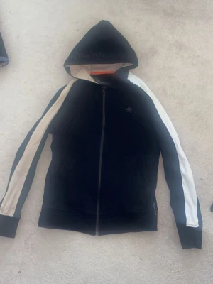 Svart hoodie med vita ränder från mooseknucles - Säljer en svart hoodie från moose knuckles med dragkedja framtill och vita ränder längs båda ärmarna. Tröjan har huva och ribbade muddar vid ärmsluten. Perfekt för dig som gillar sportig och clean stil. Materialet känns mjukt och bekvämt.