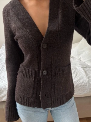 Mörkbrun stickad kofta med fickor - Mysig mörkbrun stickad kofta med tre stora knappar framtill och djupa fickor. Koftan har ribbade muddar vid ärmslut och nederkant samt en djup V-ringning. Perfekt att slänga över en t-shirt eller topp för en avslappnad look.