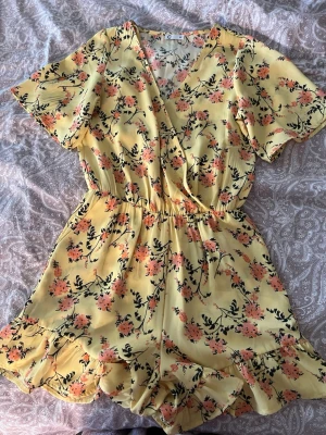 Gul blommig kort jumpsuit - Supersöt gul jumpsuit med blommigt mönster i rosa och svart. Modellen har korta ärmar, omlott-ringning och resår i midjan för en skön passform. Volangdetaljer vid benslut ger en extra touch!