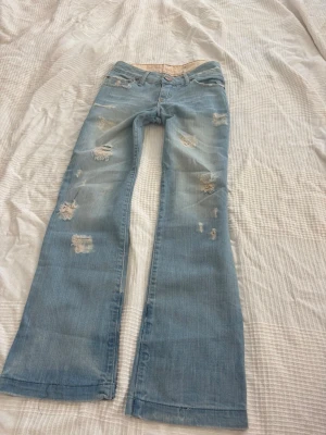 Blå slitna lowaist bootcutjeans Blanco Jeans - Säljer ett par ljusblå bootcutjeans från Blanco Jeans med slitningar och hål för en trendig, avslappnad look. Jeansen har klassisk femficksmodell, låg midja och slitna detaljer framtill och på benen. Perfekta för dig som gillar vintagevibbar och streetstyle.