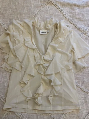 Krämvit volangblus från Kappahl - Supersöt krämvit blus från Kappahl med djup V-ringning och stora volanger framtill och på ärmarna. Blusen är tillverkad i ett tunt, lätt tyg som ger en luftig känsla. Perfekt för dig som gillar romantisk och trendig stil.