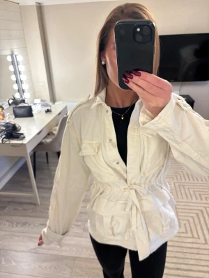 Vårjacka  - Säljer en vit vårjacka med knyt i midjan och stora bröstfickor. Jackan har klassisk krage, knappar framtill och långa ärmar. Perfekt för lager-på-lager och ger en avslappnad men snygg look.