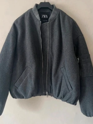 Grå slutsåld bomberjacka från Zara!! - Snygg mörkgrå bomberjacka från Zara helt slutsåld! Jackan har ribbade muddar, dragkedja framtill, två snedställda fickor och en ärmficka med dragkedja. Tillverkad i ett tjockare tyg som ger en cool och trendig look. Knappt använd så som ny🥰