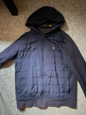 Mörkblå hoodie-cardigan från Polo Ralph Lauren - Mörkblå quiltad hoodie-cardigan från Polo Ralph Lauren med dragkedja framtill, huva med dragsko och broderad logga på bröstet. Ärmar och sidor i mjukt tyg, medan framsidan är vadderad för extra värme. Inga skador, endast nån enstaka tråd som sticker ut på ena armbågen