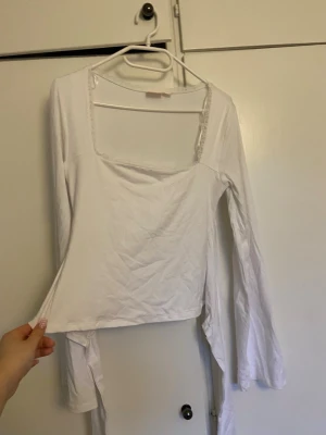 Vit långärmad topp med öppen rygg  - Trendig vit långärmad topp med fyrkantig ringning och snygg öppen rygg som knyts baktill. Tillverkad i mjukt bomullsmaterial, perfekt för dig som gillar stilrena och unika detaljer. Passar till både jeans och kjol för en cool look.