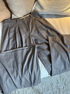 Grå vida kostymbyxor med pressveck - Säljer ett par grå/beige kostymbyxor med hög midja och vida ben. Byxorna har snygga pressveck framtill och klassiska hällor för skärp. Perfekta för en stilren och trendig look. Materialet känns mjukt och följsamt.
