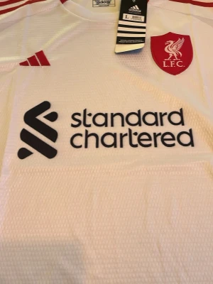 Vit Liverpool matchtröja Adidas - Snygg vit Liverpool FC matchtröja från Adidas med rött klubbmärke och sponsorlogga på bröstet. Tröjan har röda detaljer vid axlarna och är tillverkad i ett lätt, ventilerande funktionsmaterial. Perfekt för fotboll eller träning.