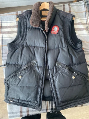 Svart dunväst från Parajumpers - Gammala dunvästen från Parajumpers Masterpiece Series med två stora framfickor med dragkedja och tryckknapp. Västen har en mjuk, brun fuskpäls på kragen och coolt patchmärke på bröstet. Perfekt för lager på lager och streetwear-vibe. 