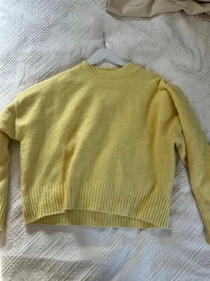 Gul stickad tröja med ribbade detaljer - Mysig gul stickad tröja med rund halsringning och breda ribbade muddar vid ärmslut och nederkant. Tröjan har en avslappnad passform och är perfekt för lager-på-lager. Materialet ser ut att vara mjukt och fluffigt, vilket ger en skön känsla.