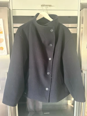 Svart ullrock med hög krage LAGER 157 💙 - Stilren svart rock i ull med rak passform och hög krage. Rocken har fem knappar framtill och vida ärmar för en modern look. Perfekt för kyliga dagar när du vill ha en enkel men snygg jacka.