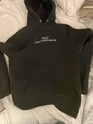 Svart hoodie från Peak Performance - Litet hål på back sidan Svart hoodie från Peak Performance med vit logga tryckt på bröstet. Klassisk modell med huva, dragsnören och känguruficka framtill. Perfekt för en avslappnad och sportig stil. Mjuk bomullsblandning som känns skön mot huden.