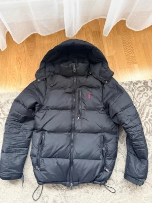Svart pufferjacka från Polo Ralph Lauren - Snygg svart pufferjacka från Polo Ralph Lauren med huva och dragkedja framtill. Jackan har två stora fickor med dragkedja och den klassiska röda loggan på bröstet. Perfekt för kalla dagar och har justerbar nederkant med snören.
