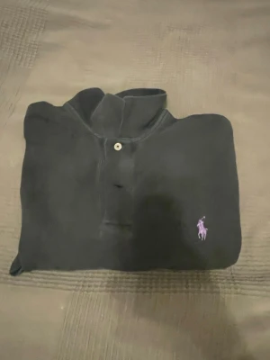 Svart pikétröja från Ralph Lauren - Klassisk svart pikétröja från Ralph Lauren med lila broderad logga på bröstet. Tröjan har krage, knappar framtill och korta ärmar. Perfekt för en clean och stilren look.