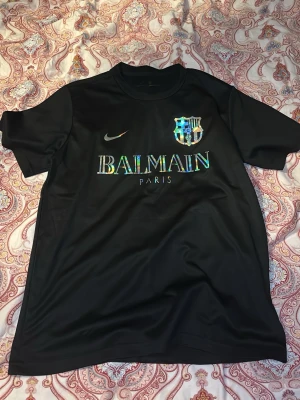 Svart Balmain x Nike t-shirt - Snygg svart t-shirt från Balmain Paris i samarbete med Nike och Barcelona. T-shirten har korta ärmar och ett holografiskt tryck med Balmain-logga och klubbmärke på bröstet. Perfekt för dig som gillar streetwear och sportig stil.