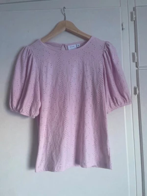 Ljusrosa blus med puffärm från Vila - Supersöt ljusrosa blus från Vila med broderade detaljer och puffiga korta ärmar. Blusen har rund halsringning och knapp i nacken. Aldrig använd.