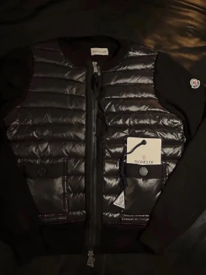 Moncler Cardigan - Snygg svart cardigan från Moncler med quiltad framsida, dragkedja och två fickor med knappstängning. Klassisk Moncler-logga på ärmen och stilren design. I storlek 1/S. Prislappen finns kvar. 