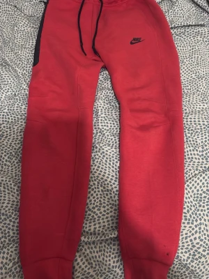 Röda Nike mjukisbyxor med svart logga - Röda mjukisbyxor från Nike med svart logga på låret och svarta dragsnören i midjan. Byxorna har muddar vid bensluten och en svart detalj vid ena fickan. Perfekta för en avslappnad och sportig stil.