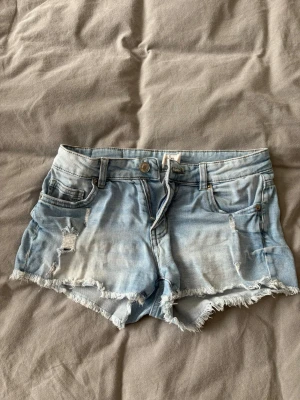 Ljusblå slitna jeansshorts - Säljer ett par ljusblå jeansshorts med fransig kant och slitna detaljer. Mid/low waisted, Perfekta för varma dagar och ger en avslappnad vibe.